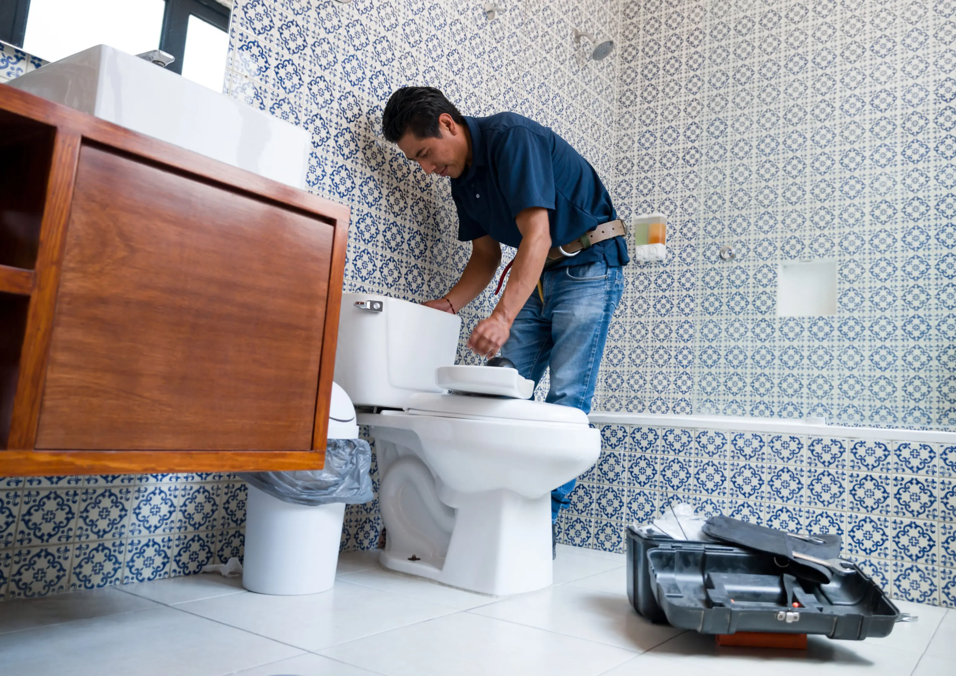 A man fixing a toilet in a tiled bathroom.