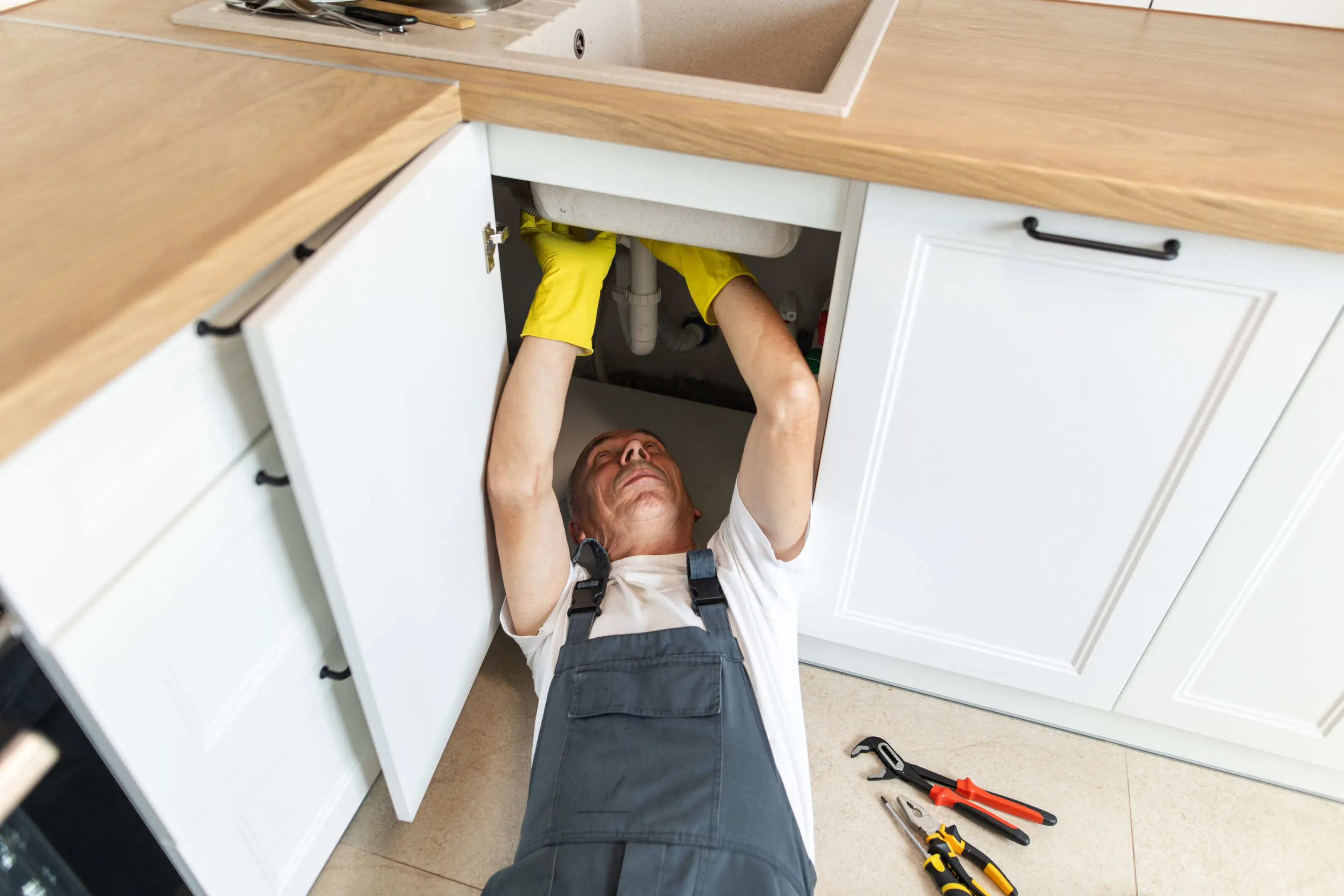 A plumber fixing pipes under a kitchen sink wearing yellow gloves.