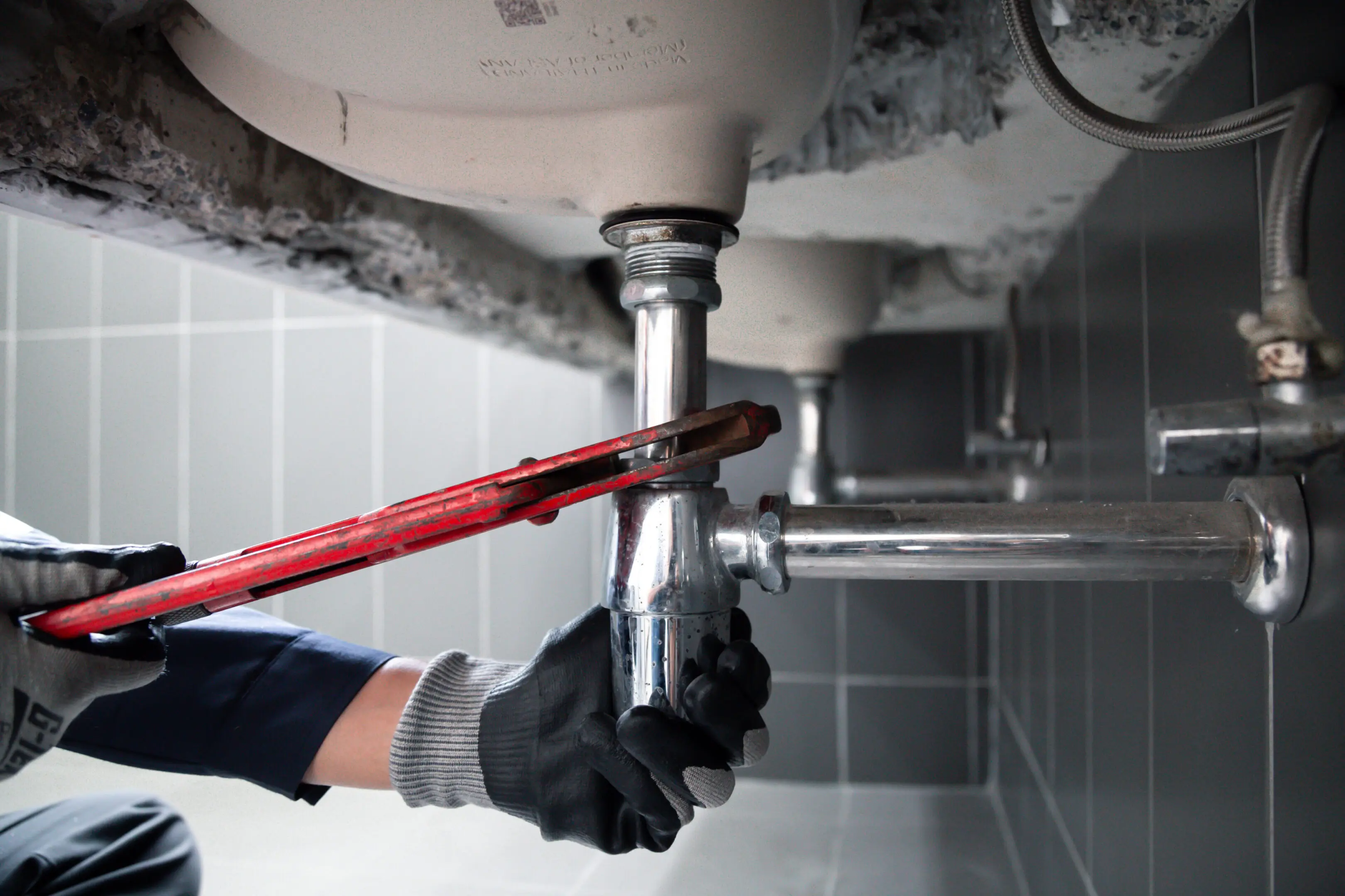 A plumber using a pipe wrench to fix bathroom plumbing under a sink.