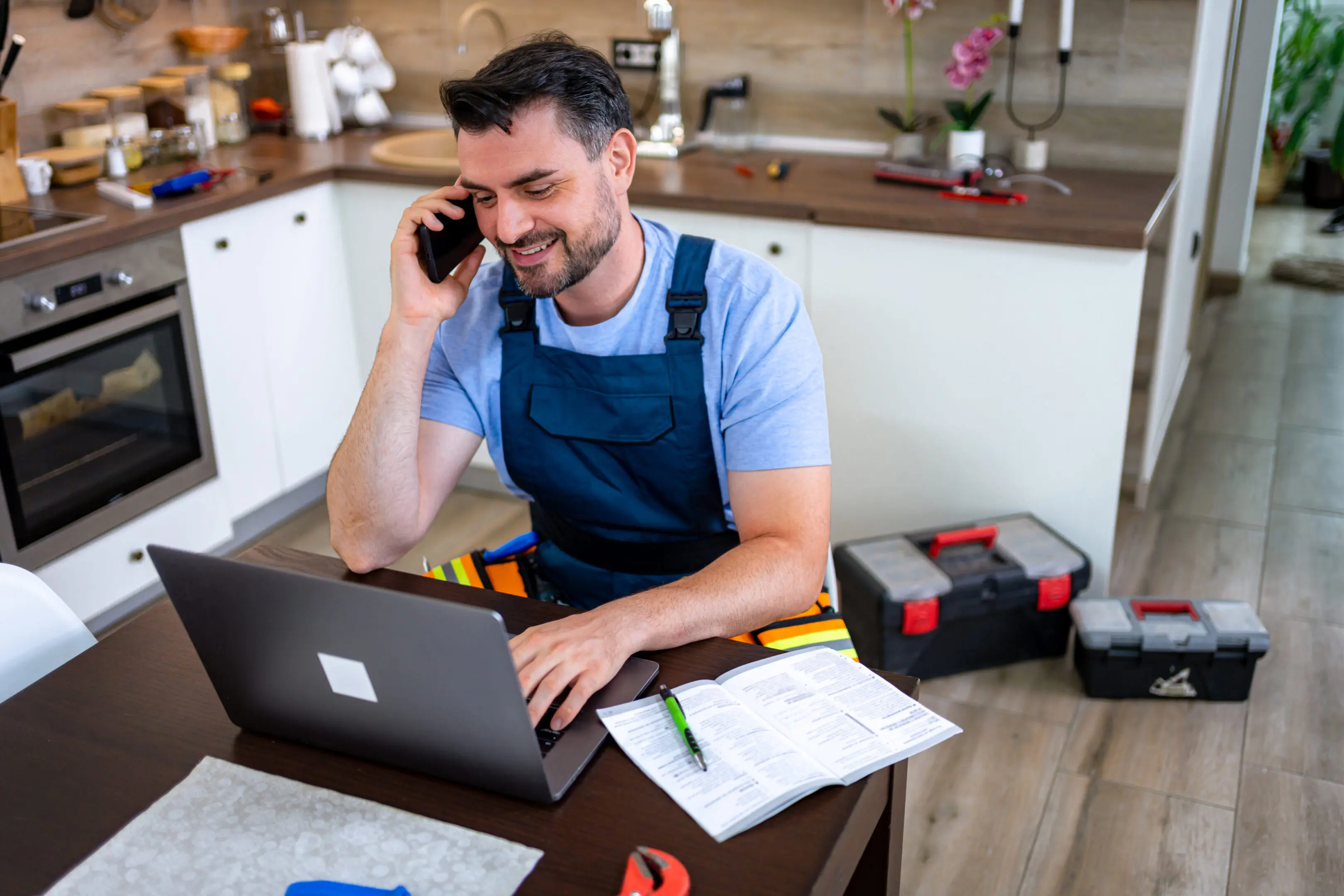 Handyman on a call while working on a laptop in a kitchen.