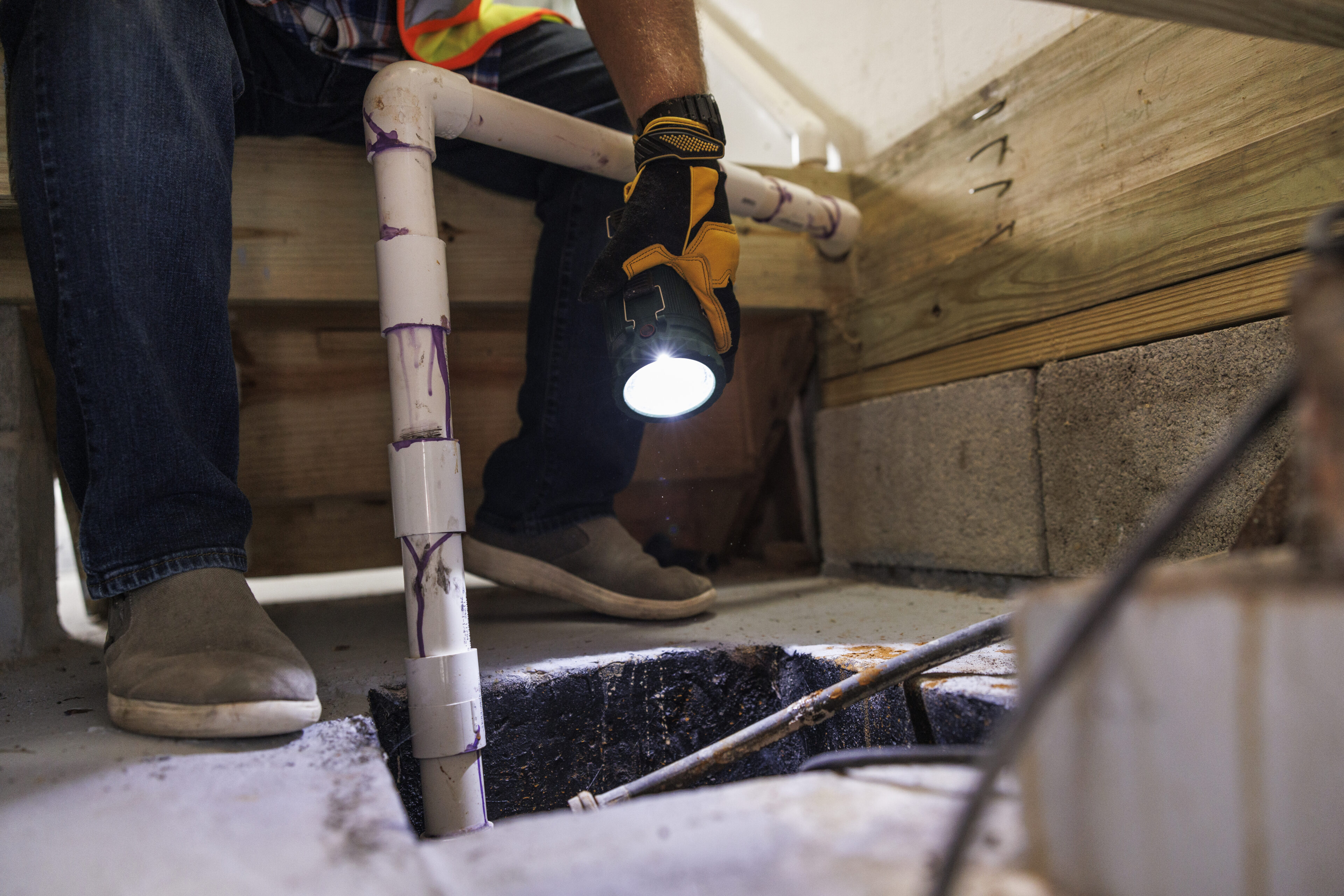 Person inspecting plumbing under a floor with a flashlight.
