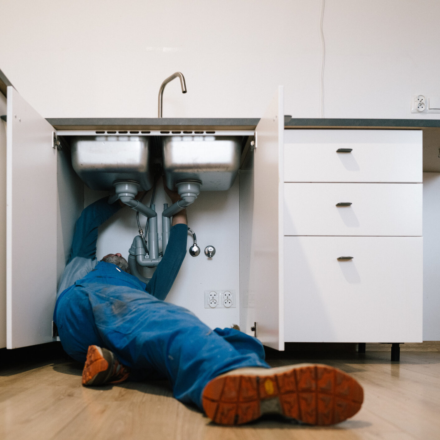 Plumber fixing pipes under kitchen sink wearing blue overalls.